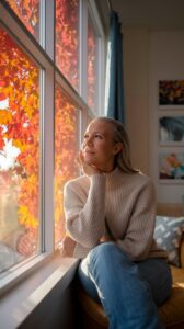 A person with long hair in a cream-colored sweater sits by a window looking outside, with orange autumn leaves visible through the glass.