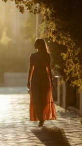 Silhouette of a woman in a long dress walking along a sunlit path lined with trees.